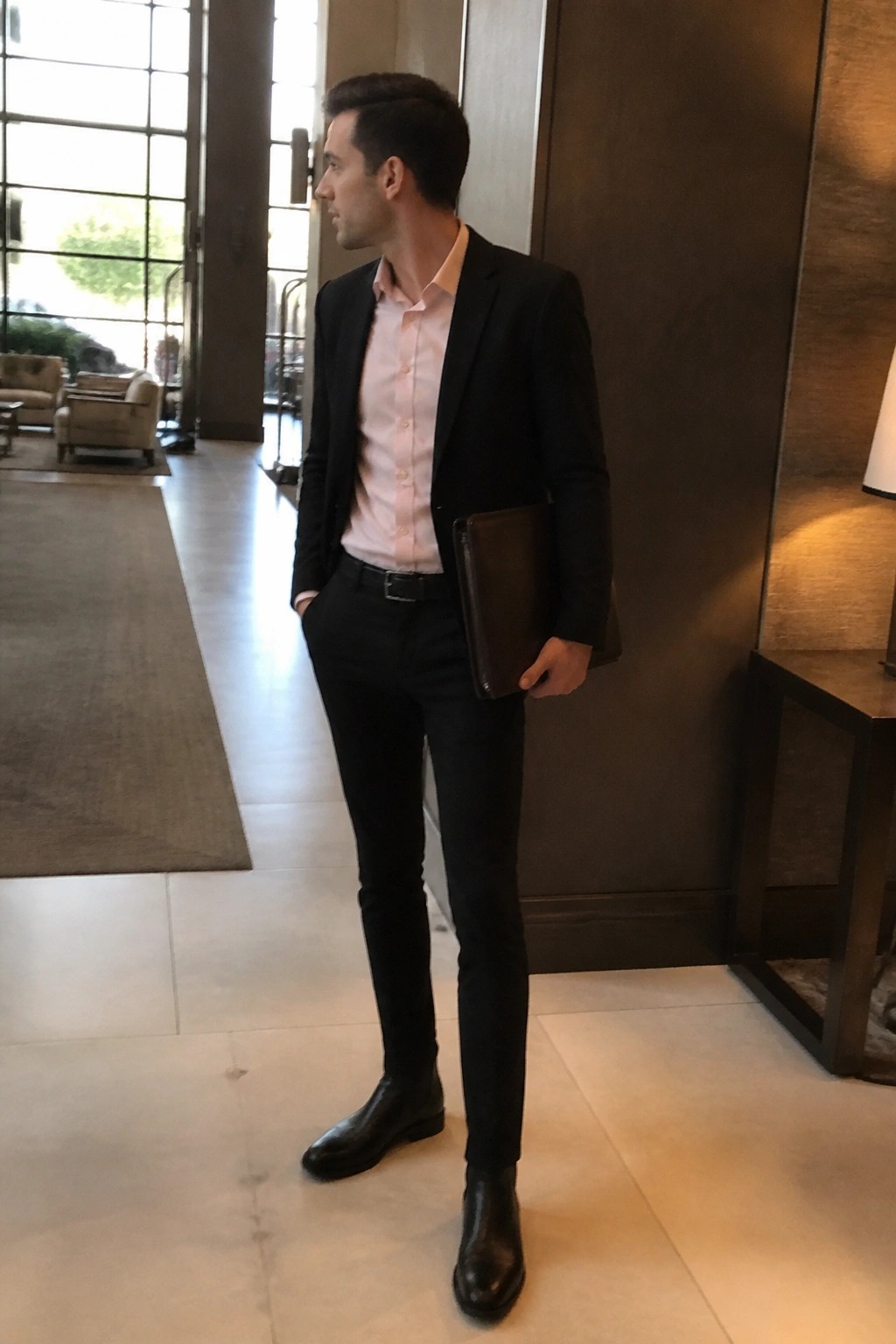 Man in black suit and pink shirt holds leather portfolio in modern lobby.