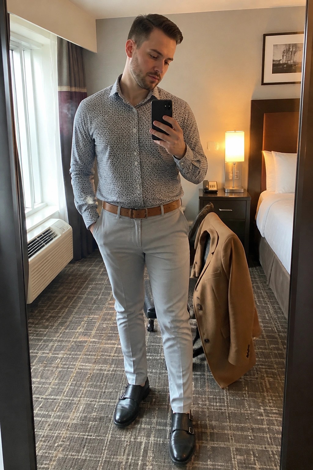 Man in patterned shirt and gray pants taking mirror selfie in hotel room