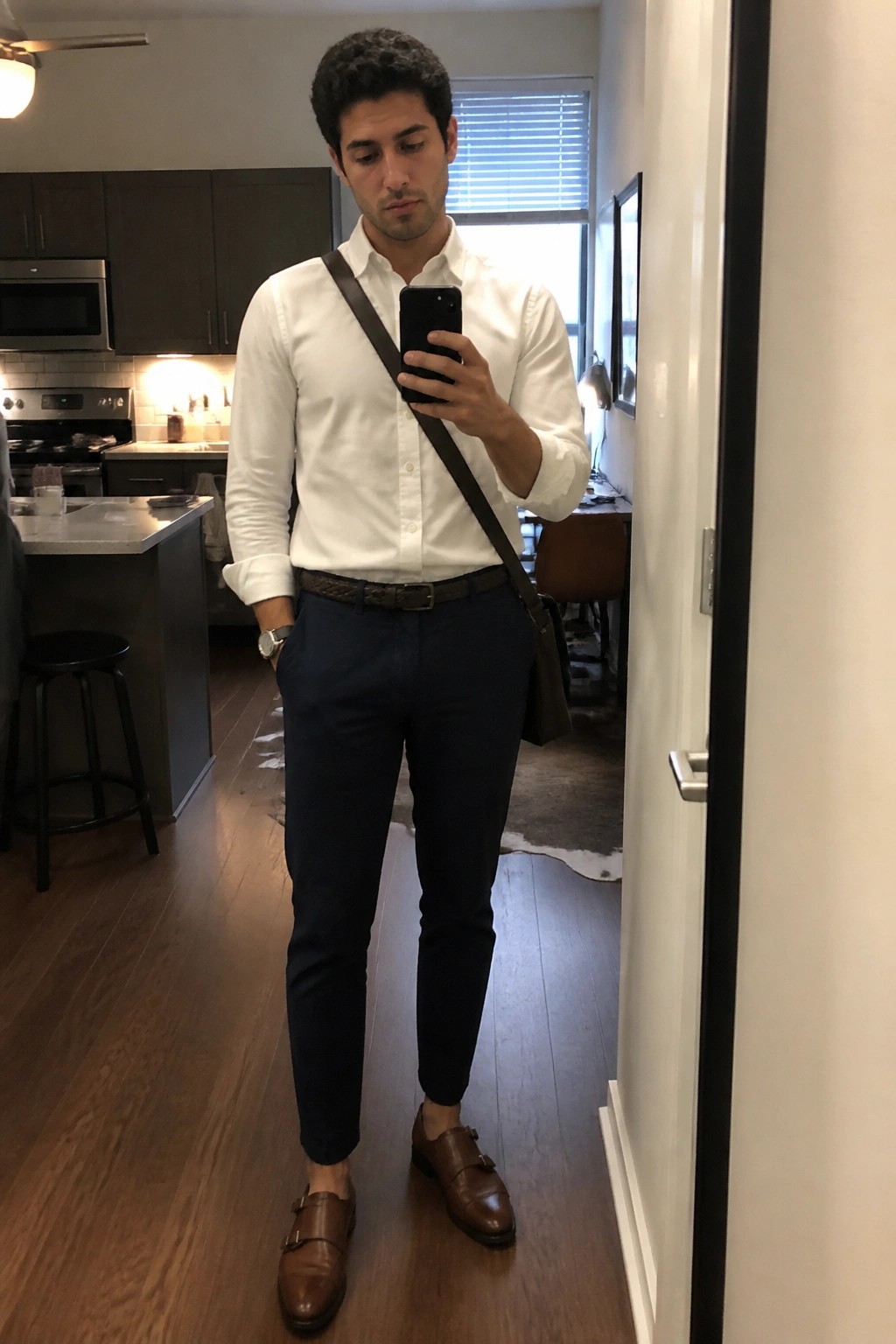 Man in white shirt and navy pants taking mirror selfie in apartment