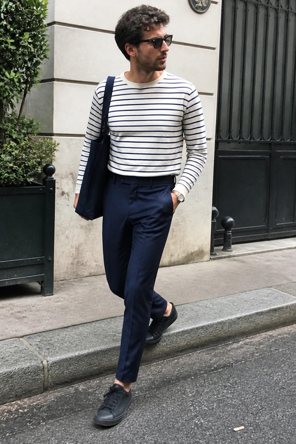 Man wearing slim navy formal pants with a white and navy striped long sleeve shirt, black sneakers, and a navy tote bag while standing on a city sidewalk.