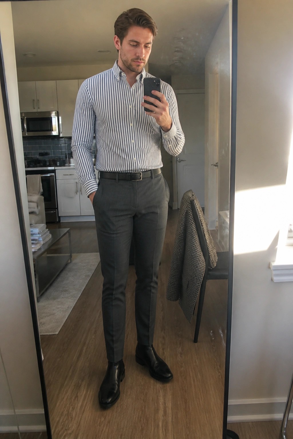 Man in striped shirt and gray pants taking mirror selfie in modern apartment