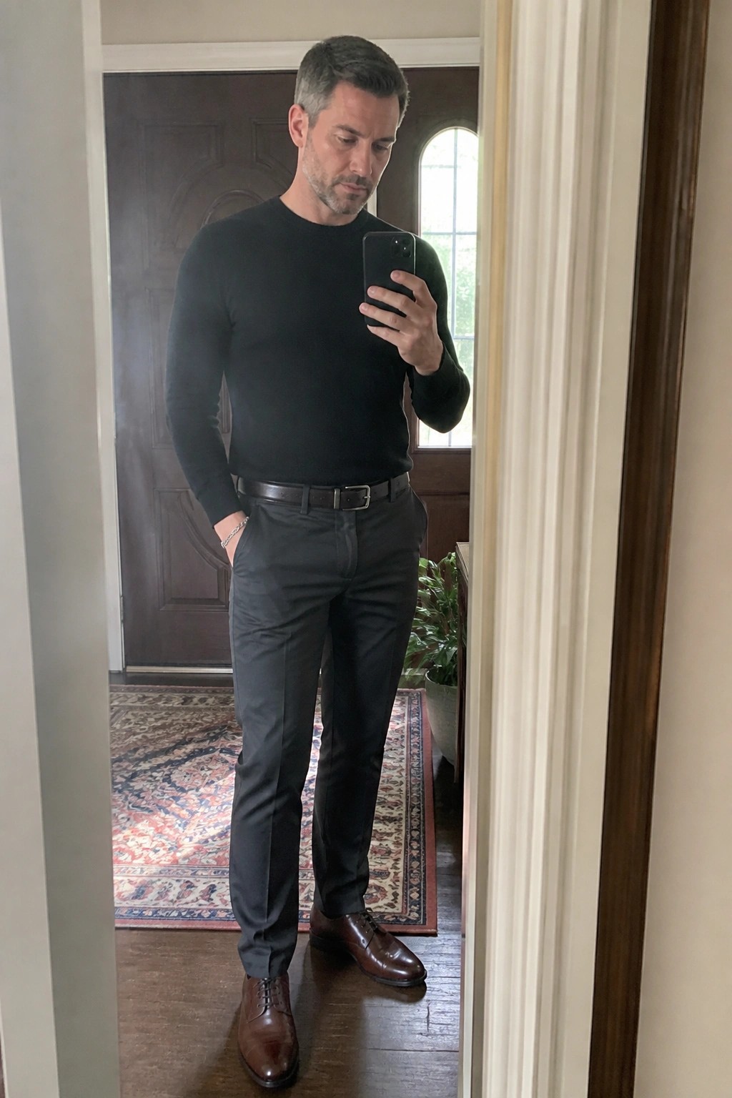 Man in slim charcoal formal pants, black long sleeve sweater, brown belt and brown dress shoes taking a mirror selfie indoors.
