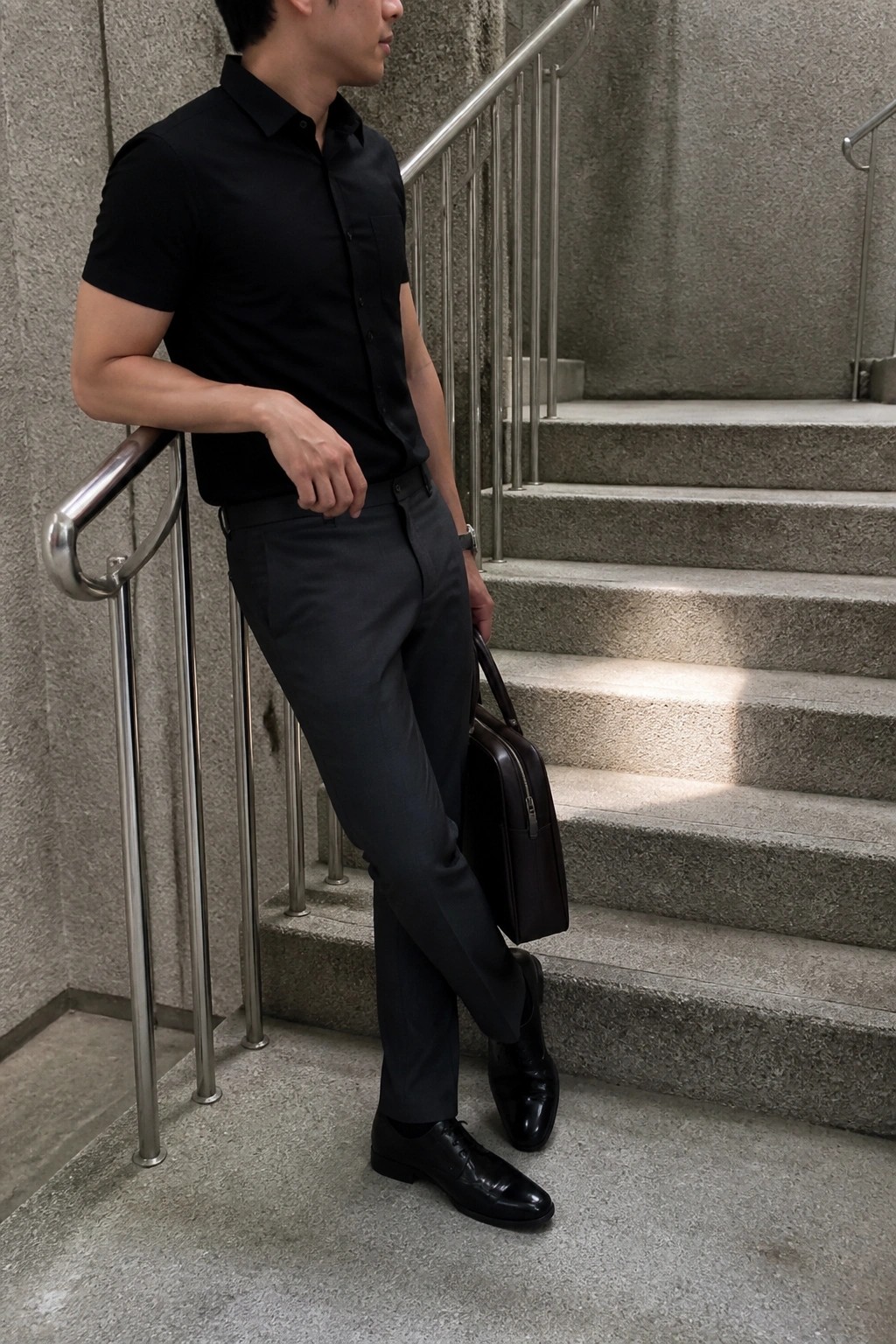 Man wearing dark gray slim fit formal pants with a black short sleeve button up shirt black dress shoes and holding a brown leather bag while standing near stairs.