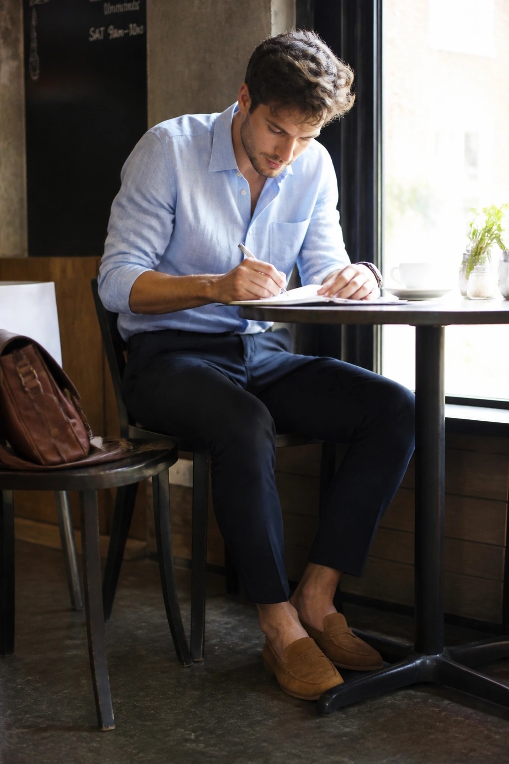 Man in light blue shirt writing at cafe table with coffee and bag.