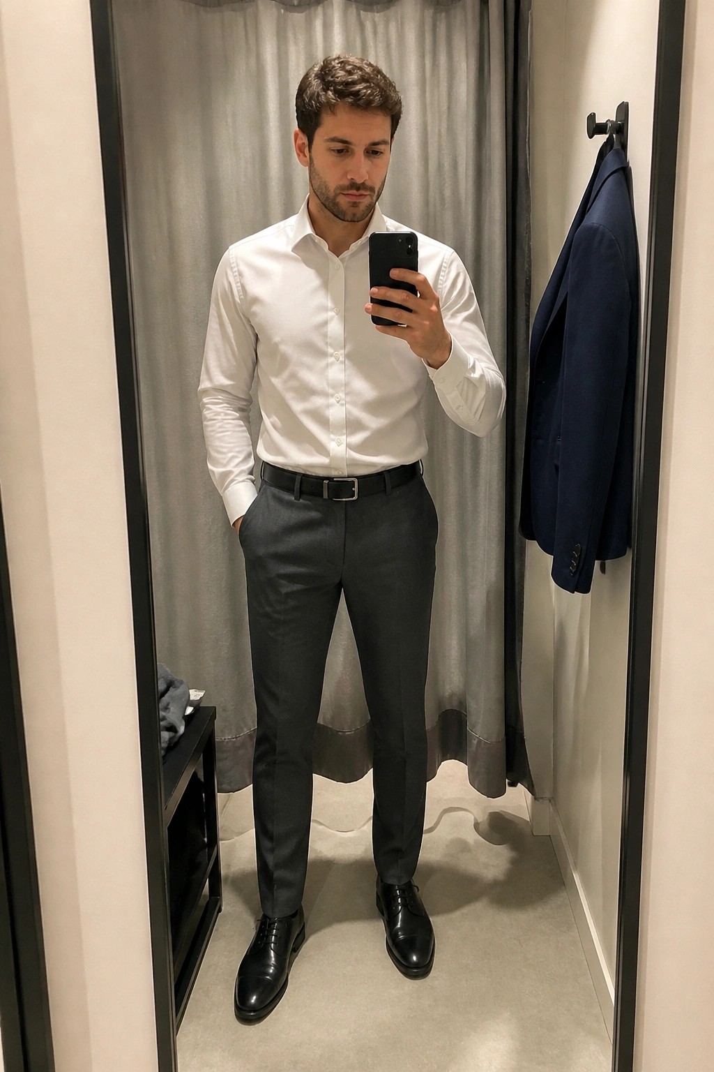 Man in slim gray formal pants, white dress shirt, black belt and black dress shoes taking a mirror selfie in a fitting room with a navy blazer hanging nearby.