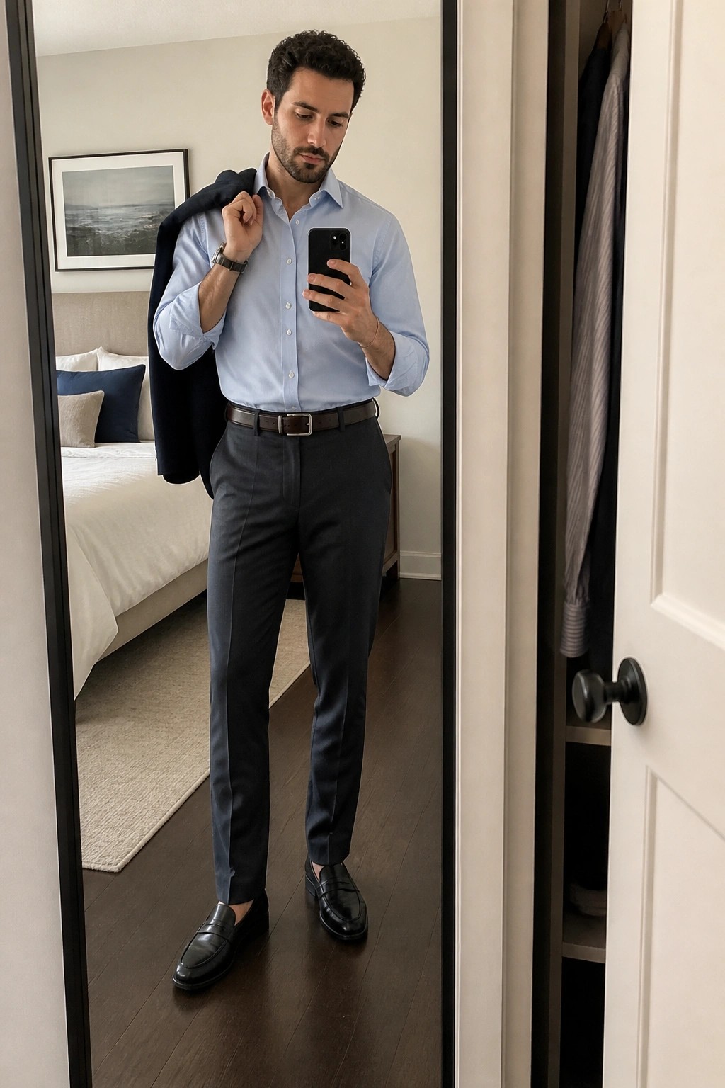 Man in light blue shirt and gray pants taking mirror selfie holding navy blazer