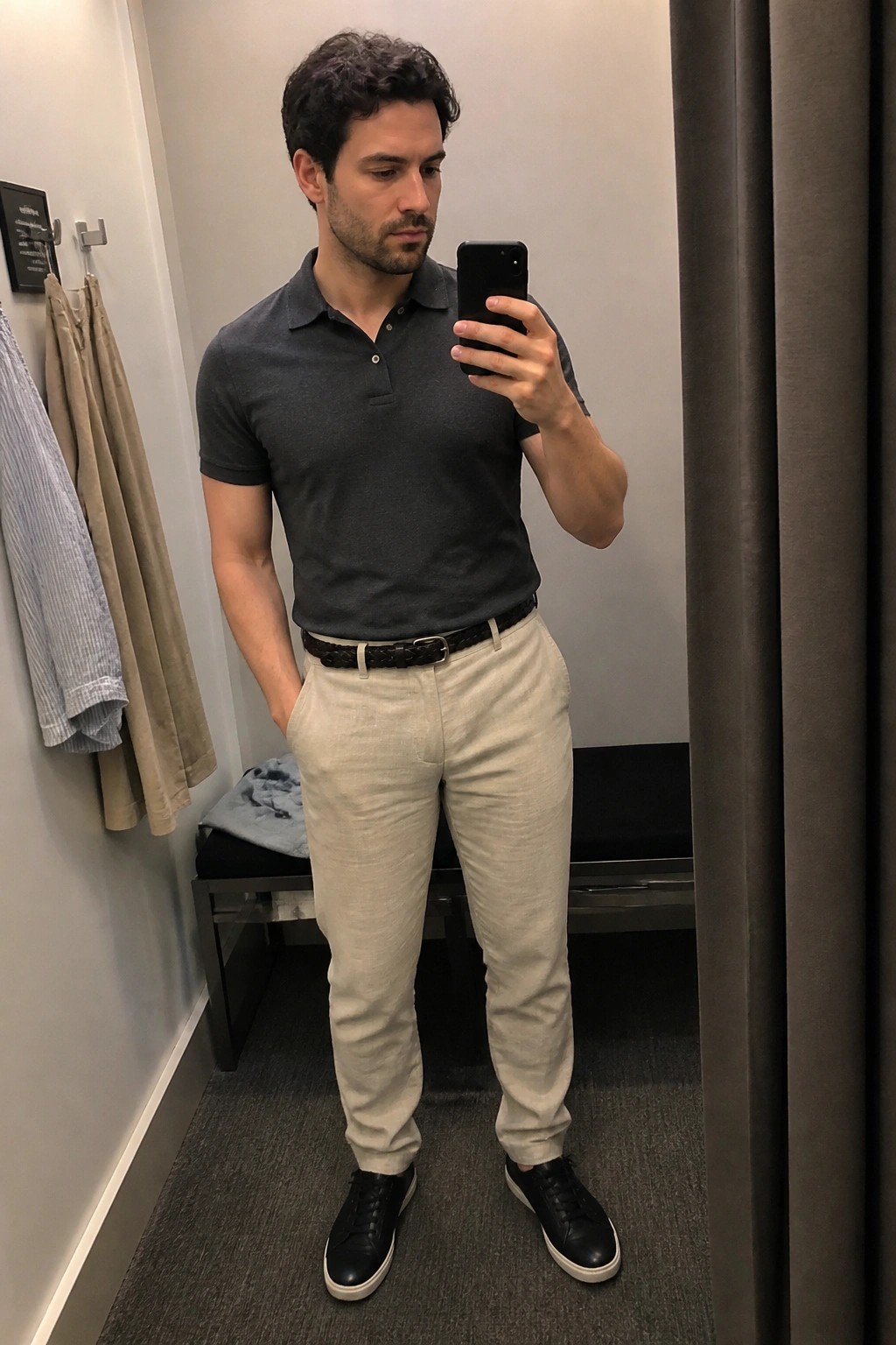 Man in gray polo shirt and beige pants taking mirror selfie in fitting room