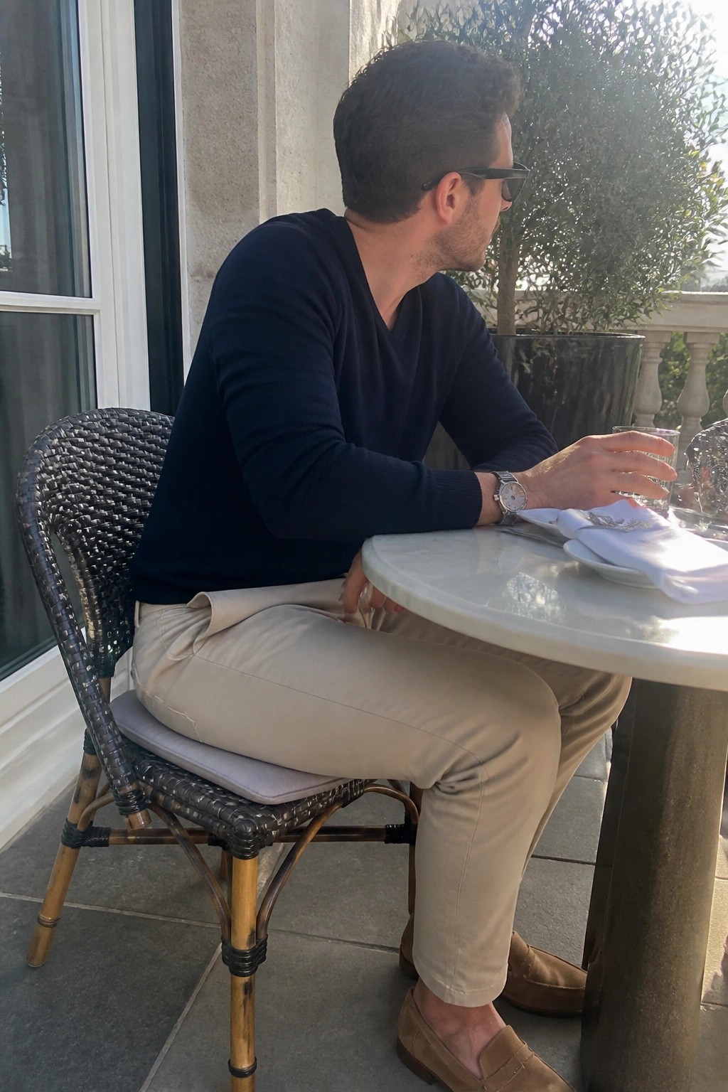 Man in navy sweater and beige pants seated at outdoor marble table.