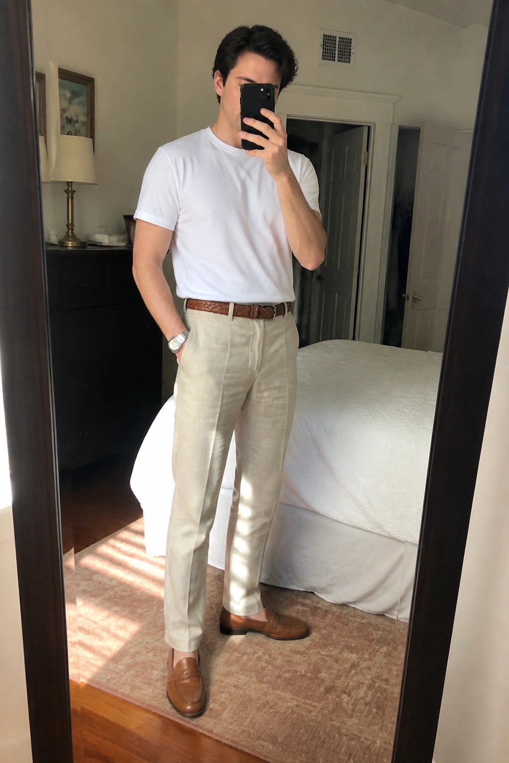 Man in white t-shirt, beige pants, brown belt and loafers taking bedroom mirror selfie