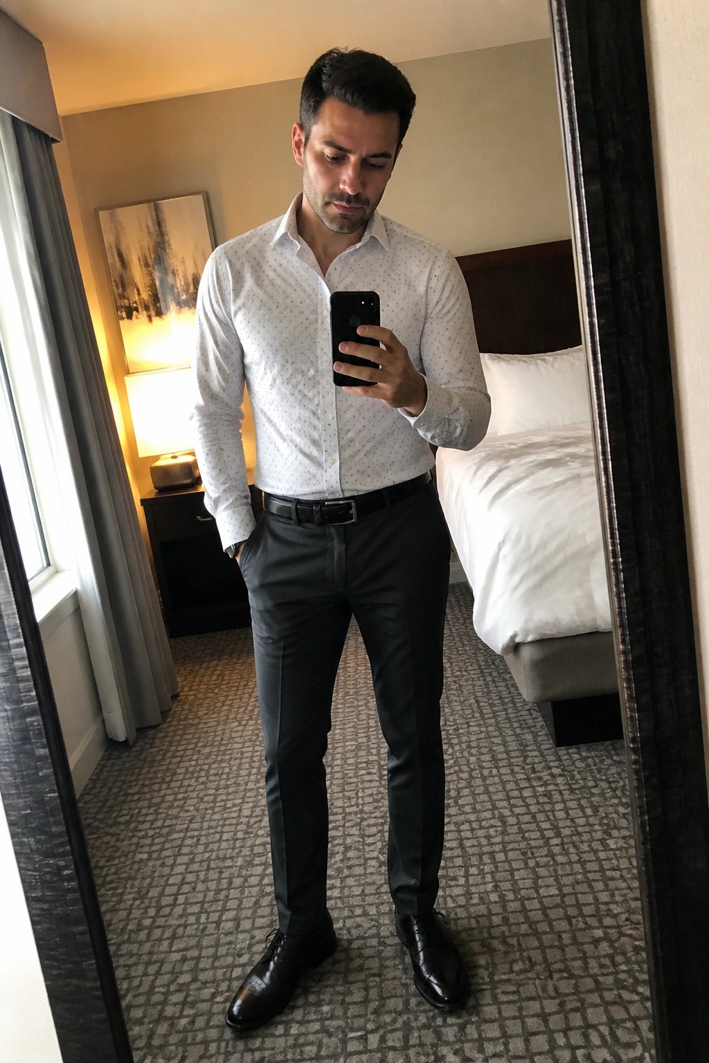 Man in white patterned button down shirt tucked into black formal pants with black belt and black dress shoes standing in a hotel room taking a mirror selfie with a phone.