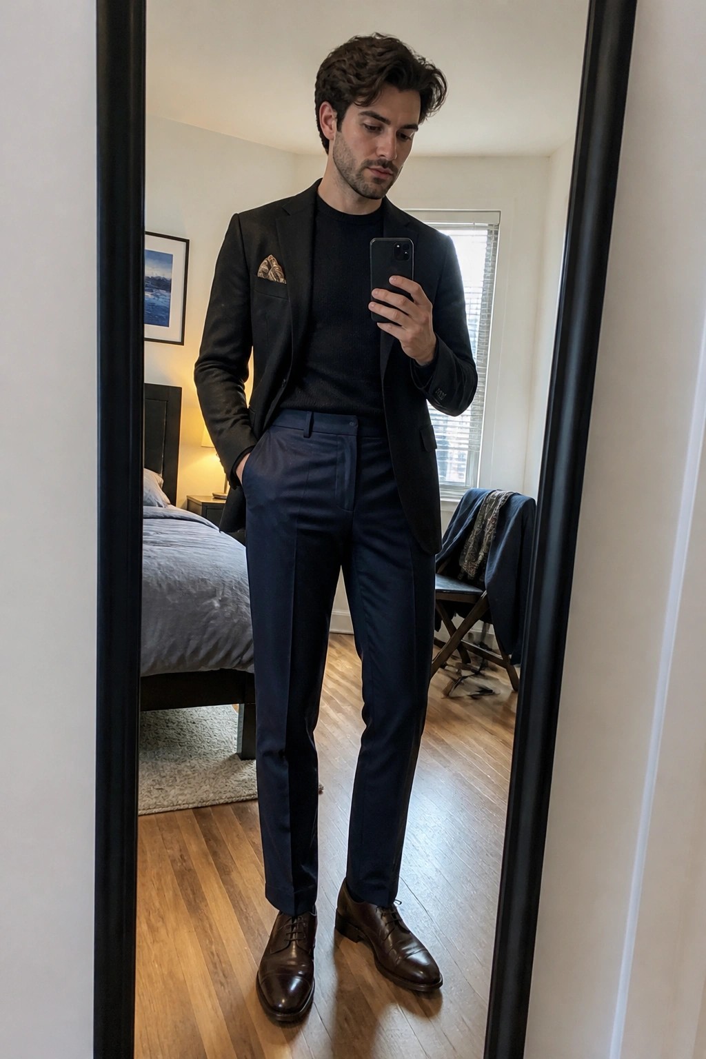 Man wearing navy formal pants, black blazer over black sweater, patterned pocket square, and brown leather dress shoes while taking a mirror selfie in a bedroom.