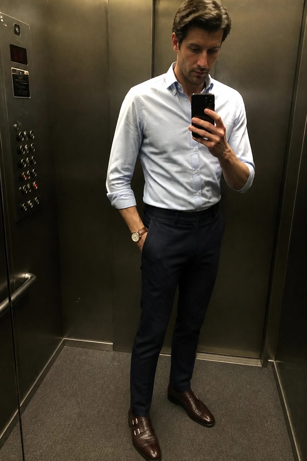 Man in light blue shirt and navy pants taking selfie in elevator