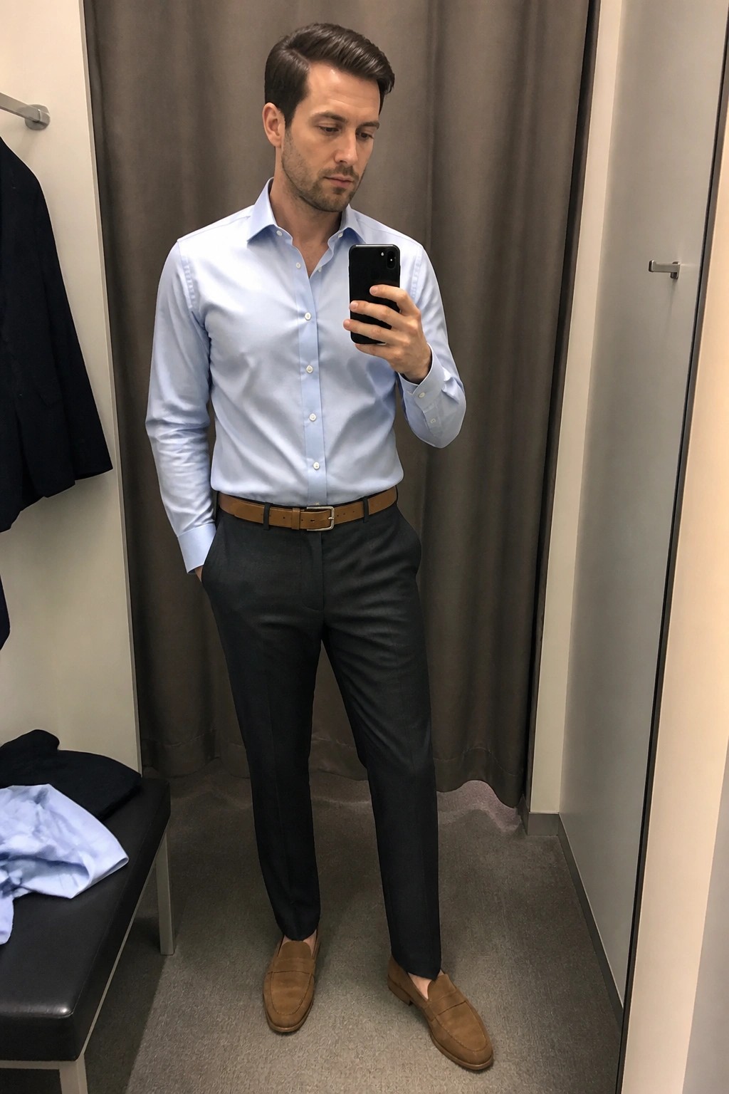Man in light blue shirt and gray pants taking mirror selfie in fitting room