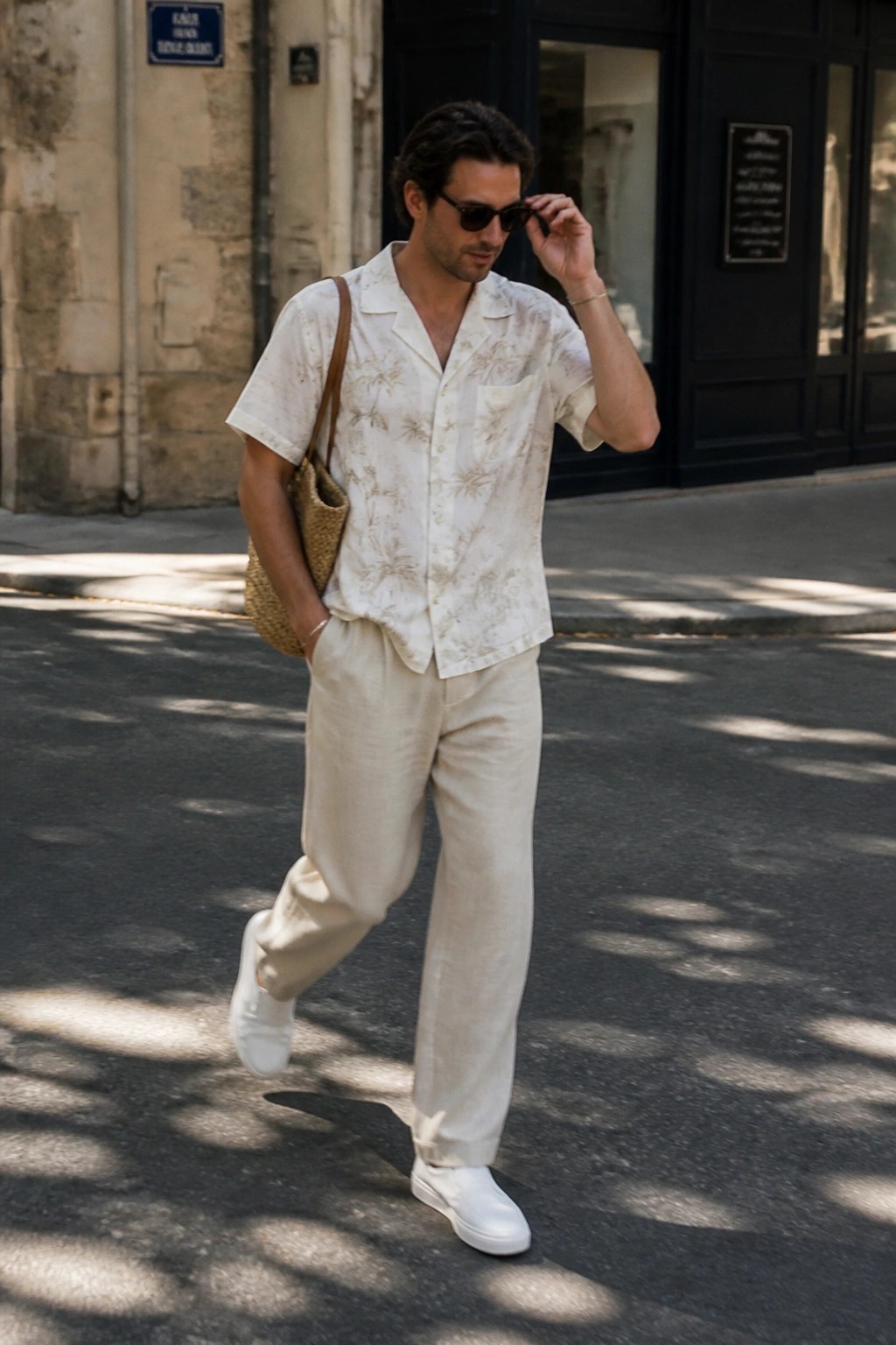 Man wearing relaxed off-white linen pants, cream short-sleeve patterned button-up shirt, white sneakers and carrying a woven shoulder bag.