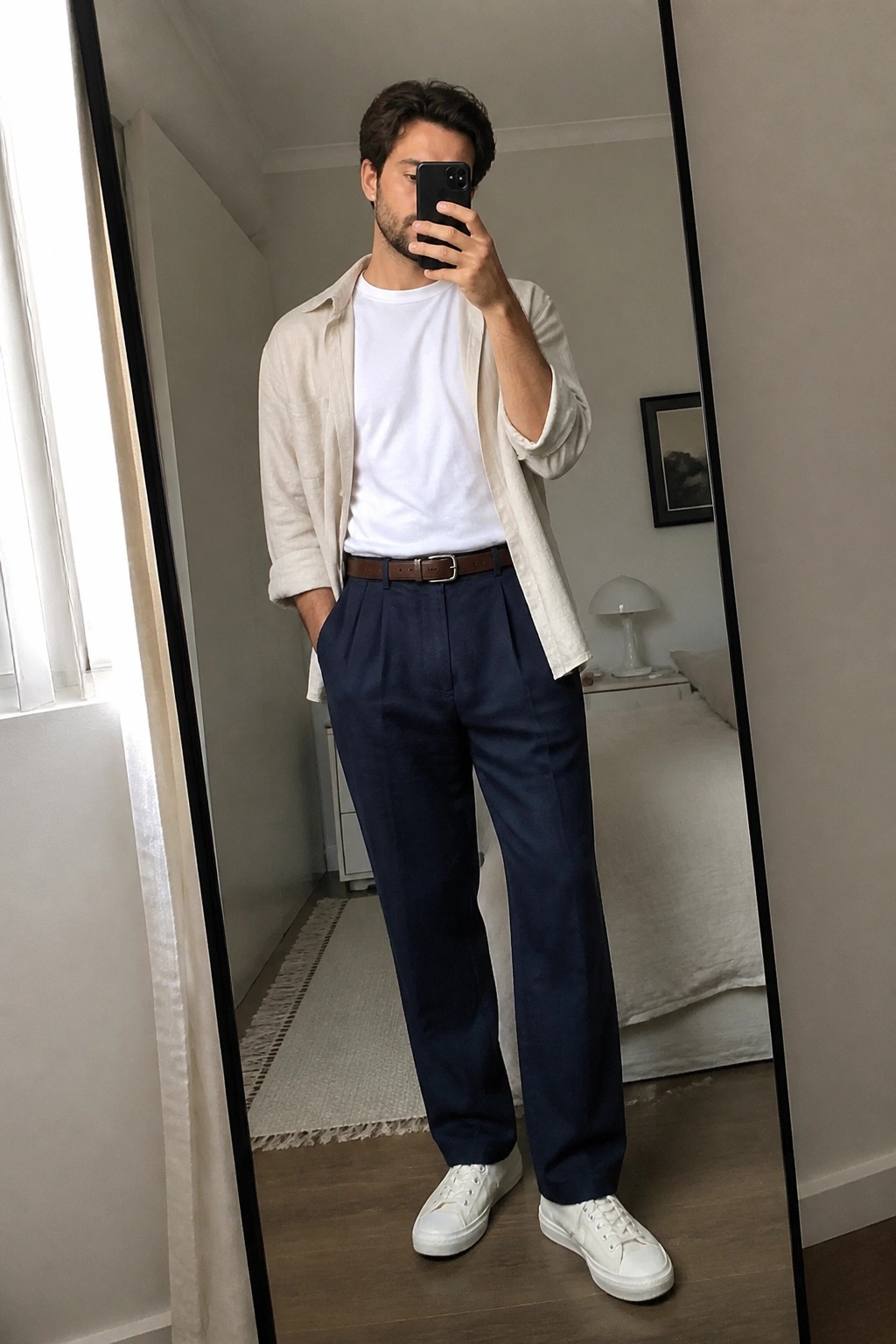Man in white tee, beige shirt, navy trousers taking mirror selfie in bedroom.
