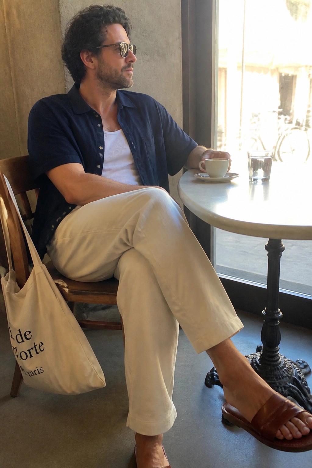 Man seated wearing off-white relaxed linen pants, navy short-sleeve button-up shirt open over a white tank top, brown leather slides, and a beige tote bag resting on the chair beside him.