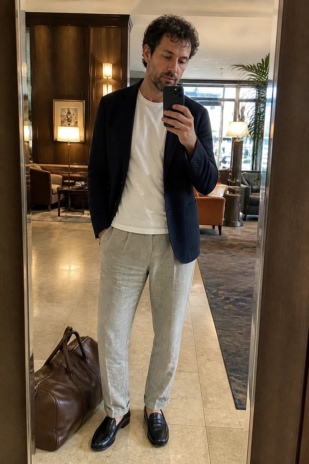 Man wearing light gray relaxed linen pants, navy blazer over white t-shirt, and black loafers while holding a phone in a mirror selfie with brown duffel bag visible nearby.
