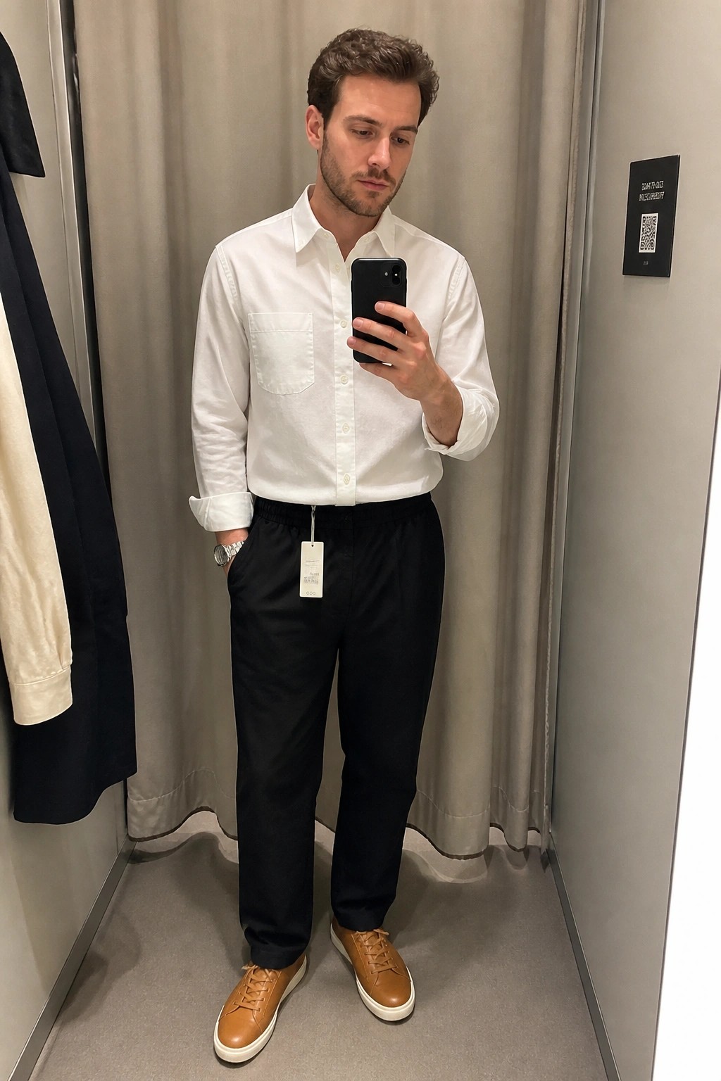 Man in white shirt and black pants taking mirror selfie in fitting room