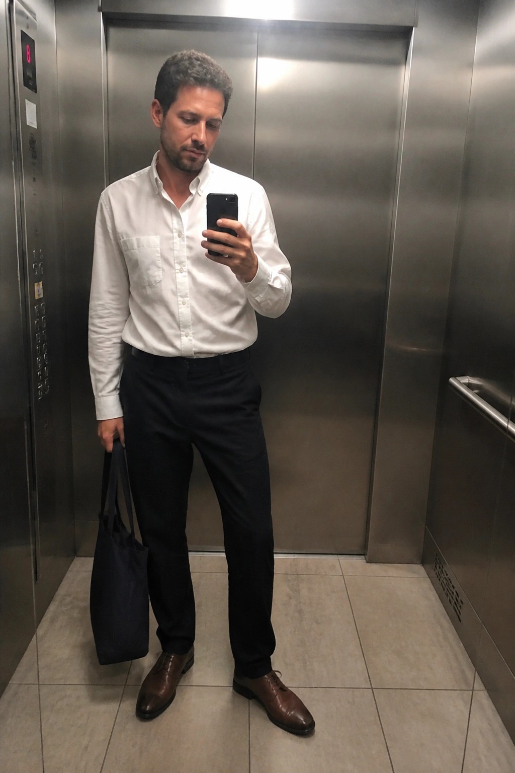 Man in white shirt and black pants taking phone selfie in elevator holding blue bag