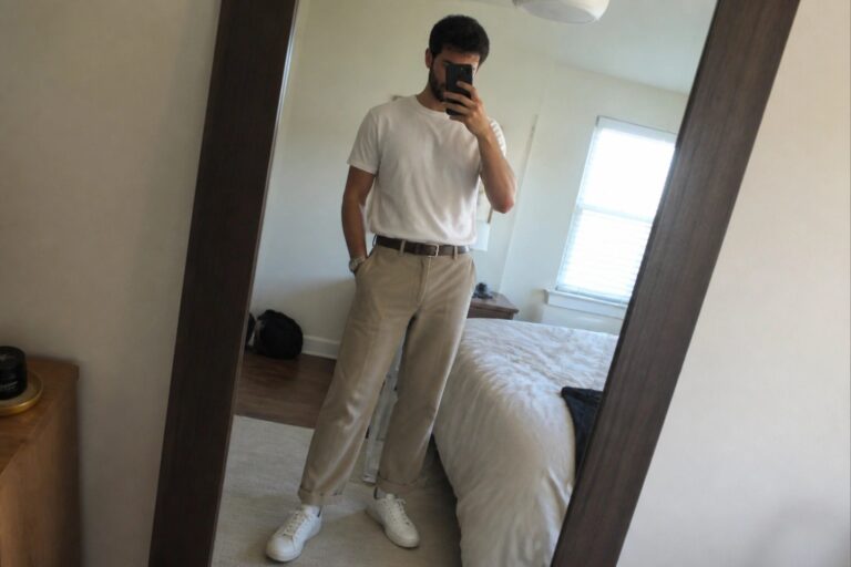 Man in white t-shirt and khaki pants takes mirror selfie in bedroom.