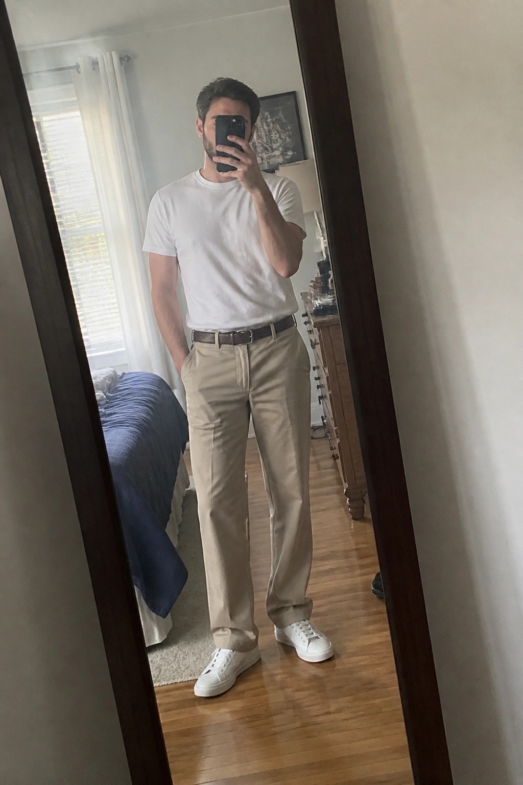 Man in white t-shirt and khaki pants takes mirror selfie in bedroom.