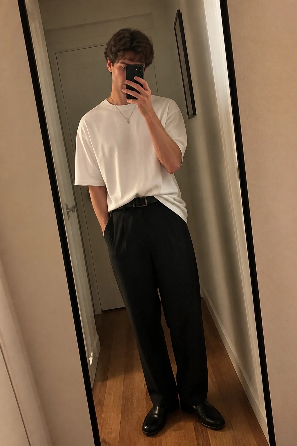 Man in loose white t shirt tucked into black relaxed straight leg pants with black belt and black loafers holding phone for mirror selfie in hallway