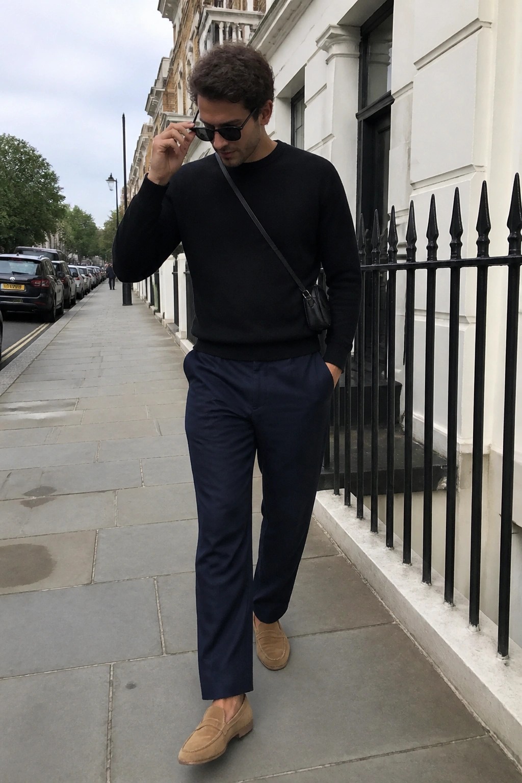 Man in black sweater and navy pants adjusting sunglasses on city sidewalk