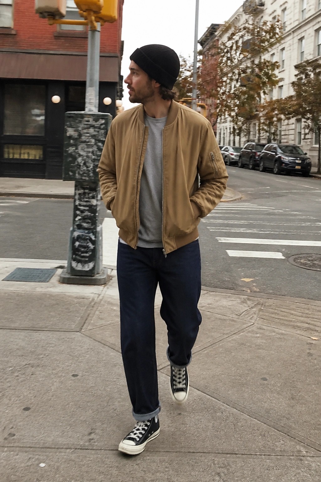 Man in tan bomber jacket and black beanie walks on city sidewalk.