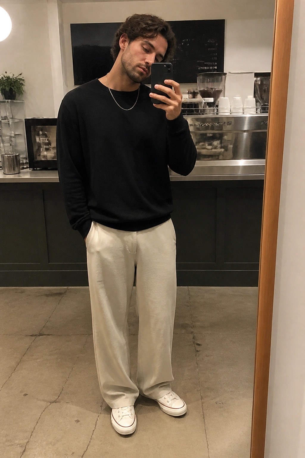 Man in black sweater and white pants taking mirror selfie in cafe