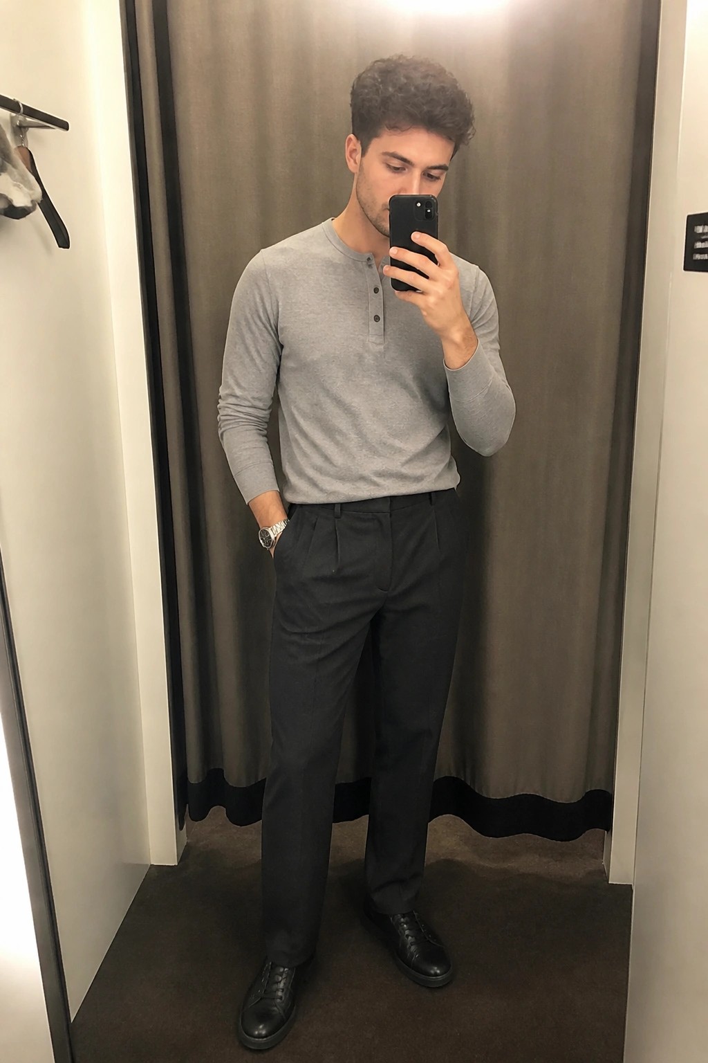 Young man in gray henley shirt and dark pants takes mirror selfie in fitting room.