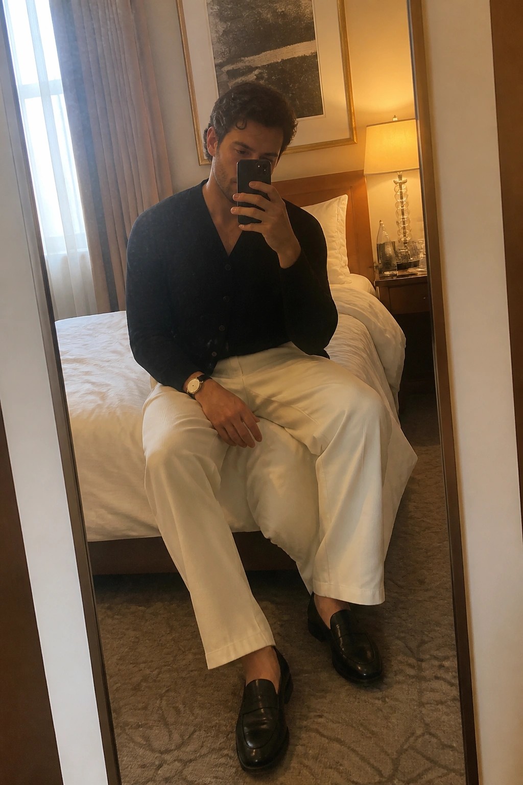 Man seated on bed wearing navy button cardigan, white relaxed straight leg pants, and black loafers while holding phone for mirror selfie.