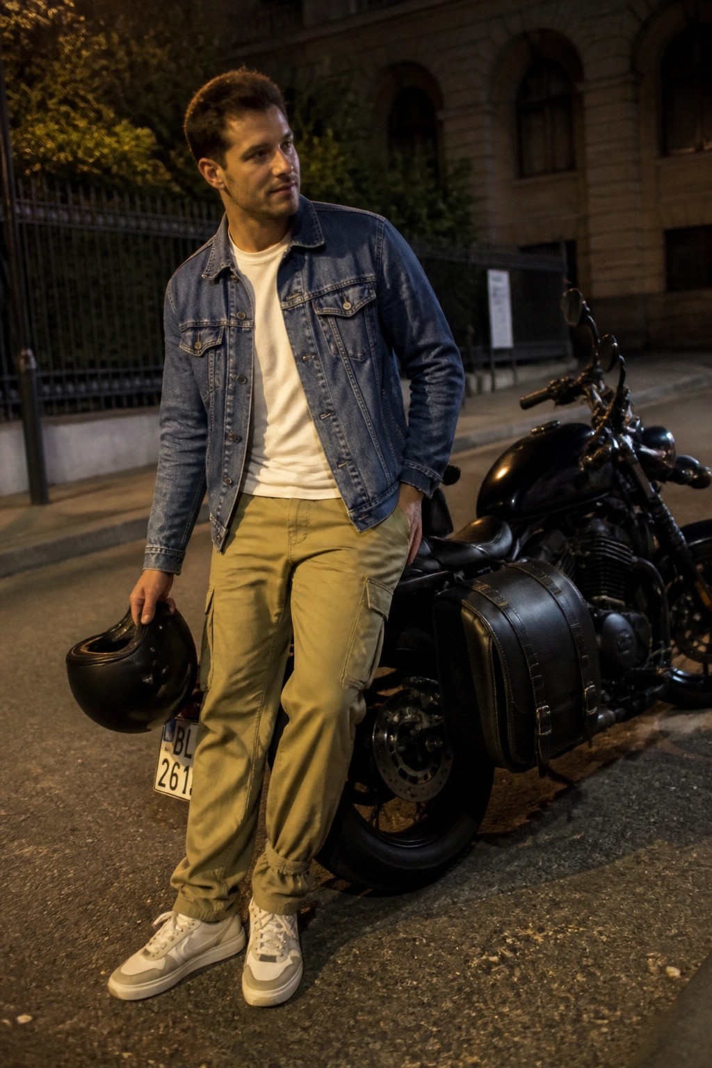 Man wearing khaki cargo pants, white t-shirt, blue denim jacket, and white sneakers while holding a black motorcycle helmet beside a black motorcycle.