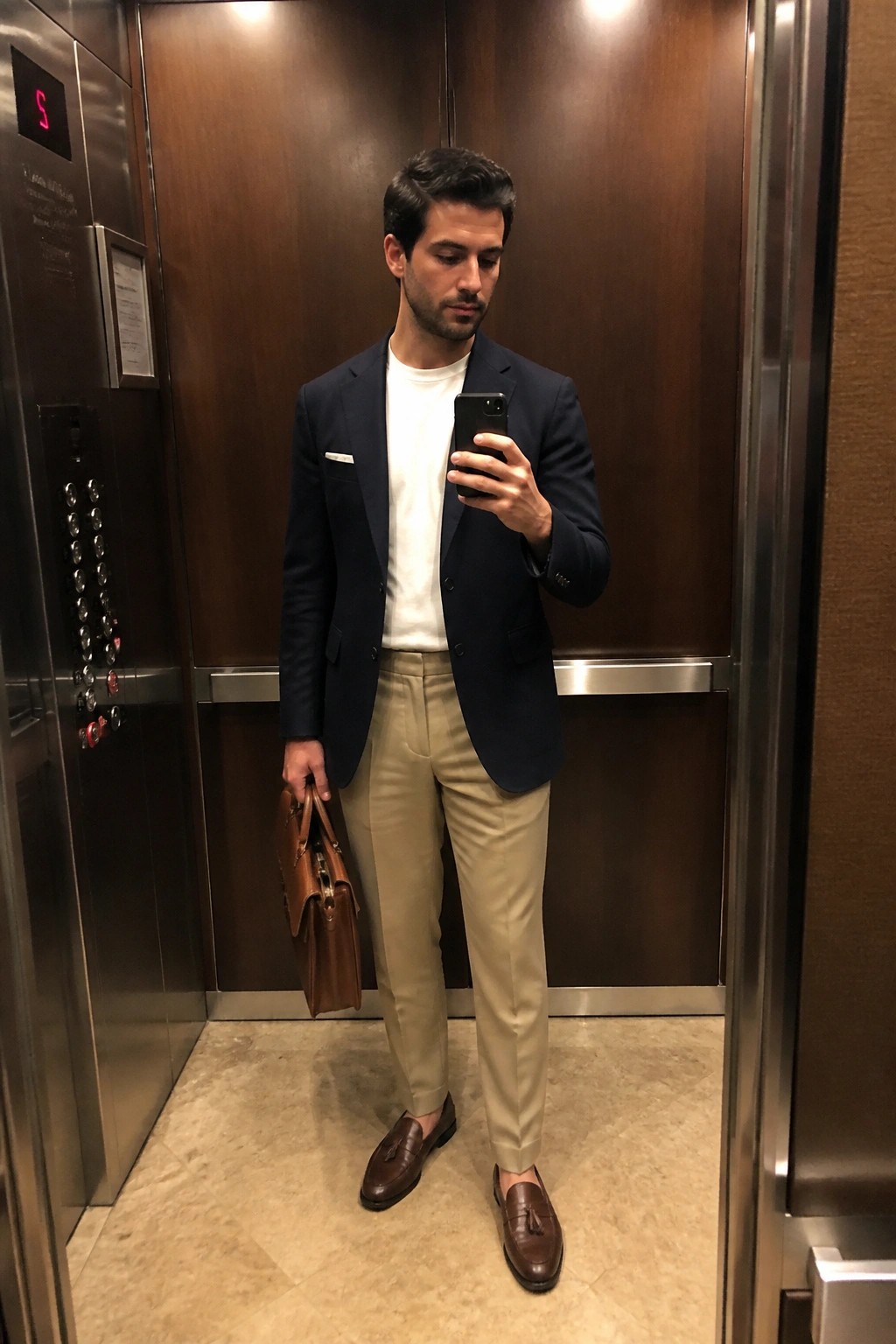 Man wearing khaki pants with white t-shirt, navy blazer, brown tassel loafers and carrying a brown leather bag while taking a mirror selfie in an elevator.