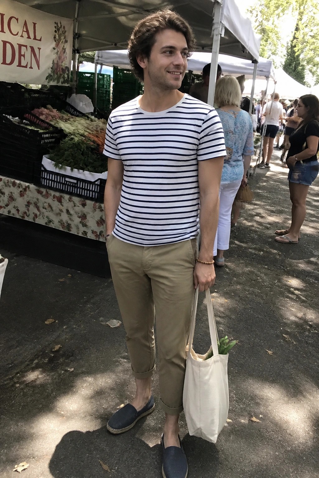Man wearing rolled khaki pants, navy striped short sleeve shirt, blue espadrilles and holding a canvas tote at an outdoor market.