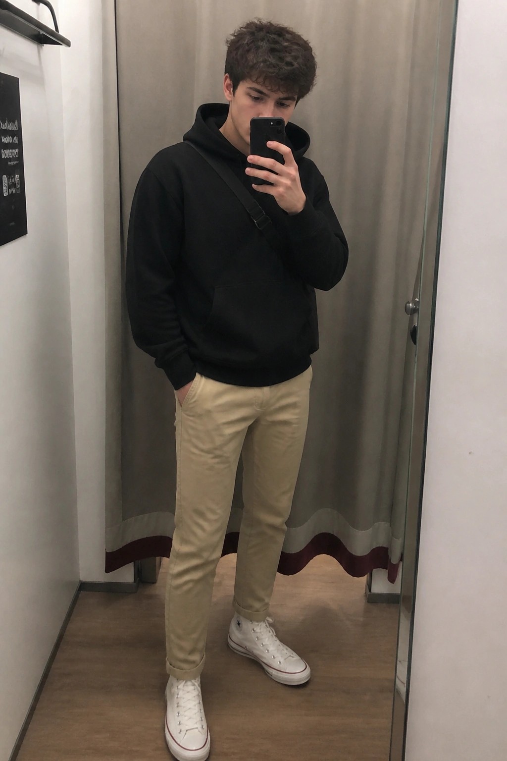 A young man in khaki pants, black hoodie, white Converse sneakers, and crossbody strap taking a mirror selfie in a hallway.