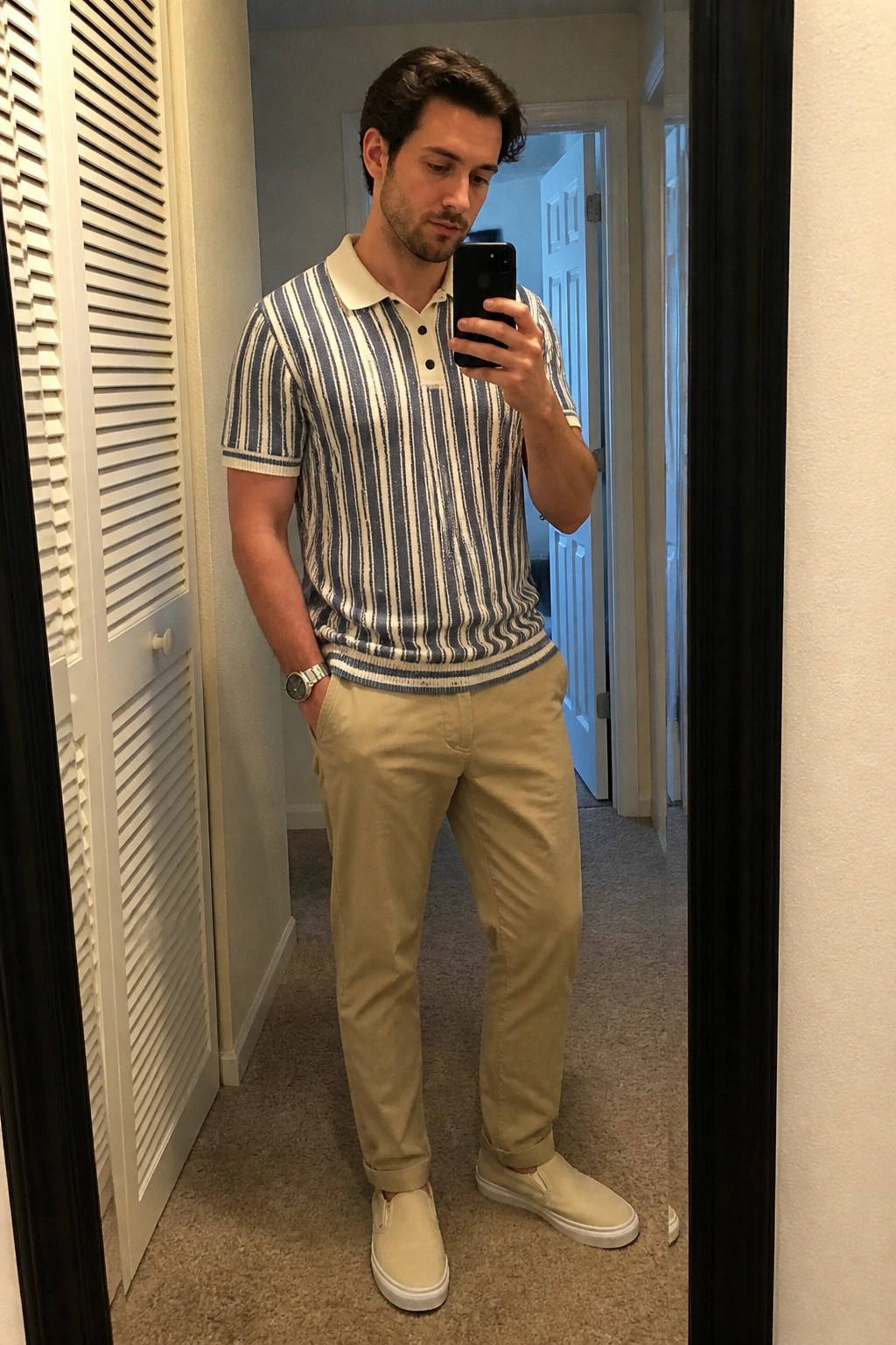 Man in striped polo shirt and khaki pants taking mirror selfie with phone.