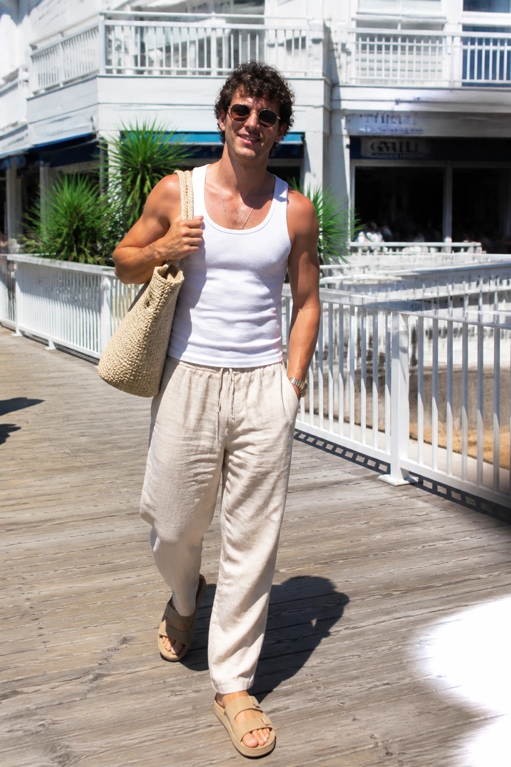 Man in white tank top and beige pants carrying woven bag on boardwalk
