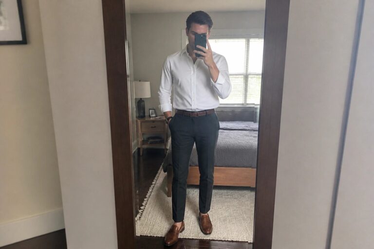 Man in white shirt and gray pants taking bedroom mirror selfie with phone.