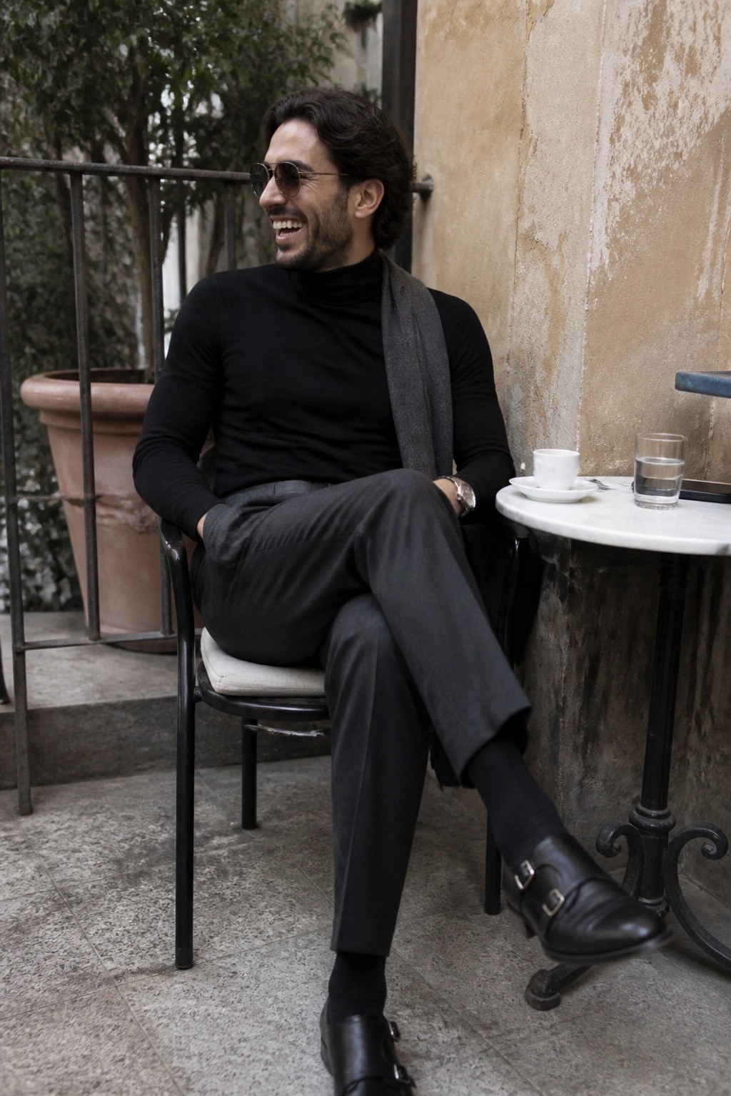 Man seated outdoors wearing black turtleneck sweater, gray tailored pants, black double monk strap shoes, gray scarf, and sunglasses in a relaxed pose.