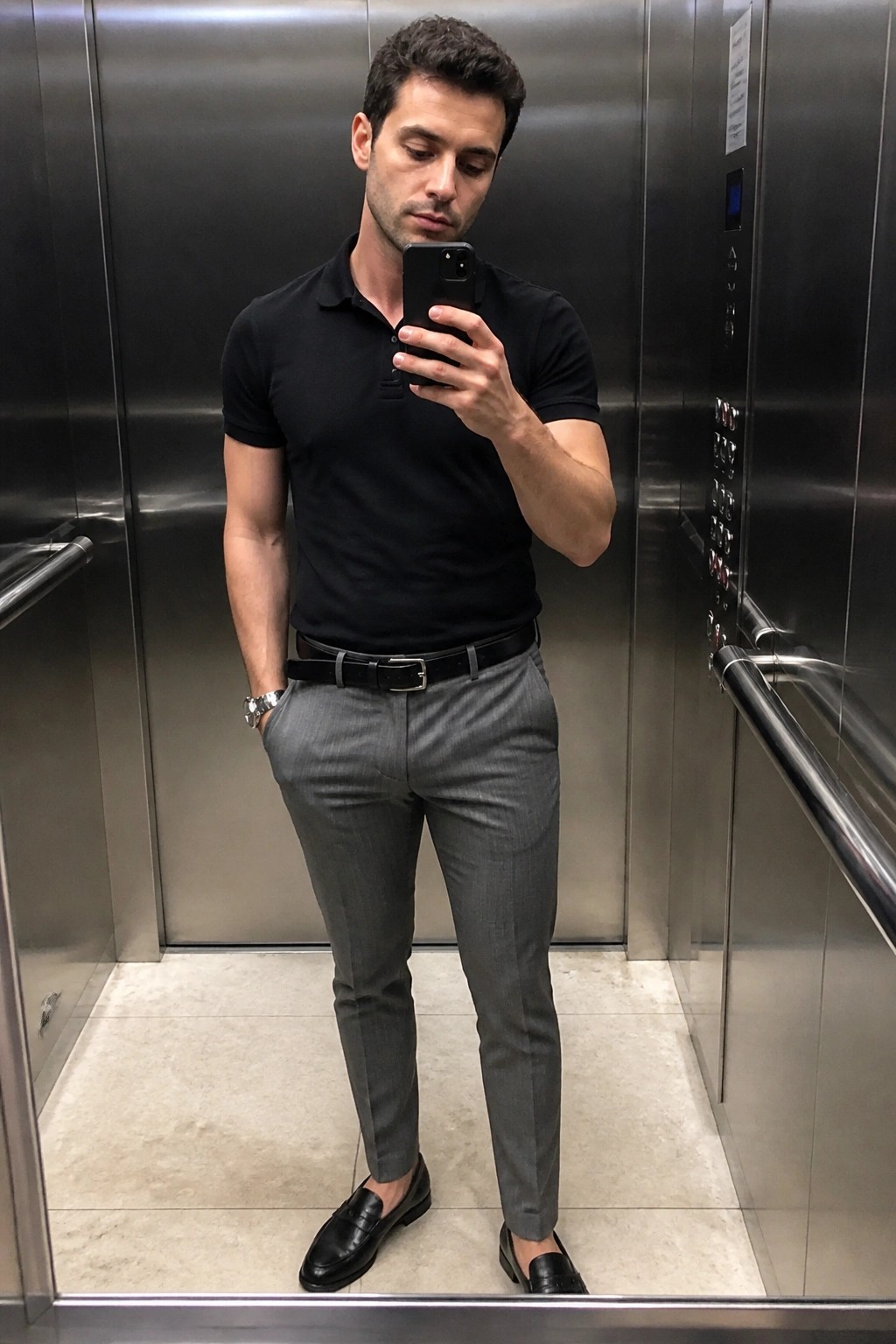Man in black polo shirt and gray pants taking mirror selfie in elevator