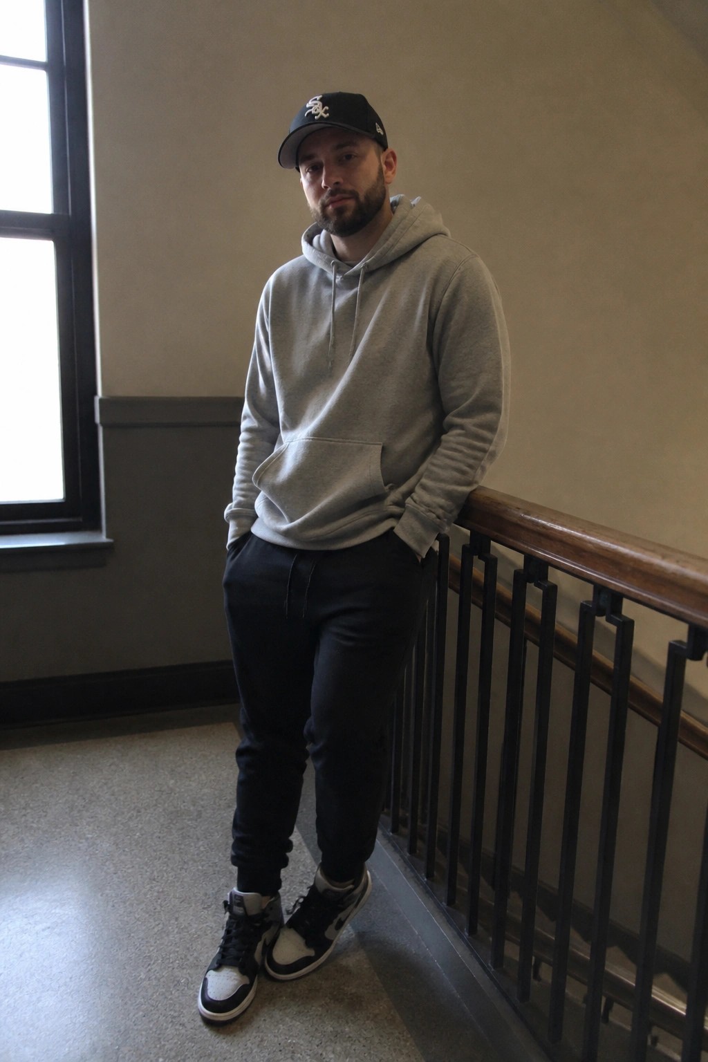 Man wearing gray hoodie, black jogger pants, and black and white high top sneakers with a black baseball cap standing by a wooden railing indoors.