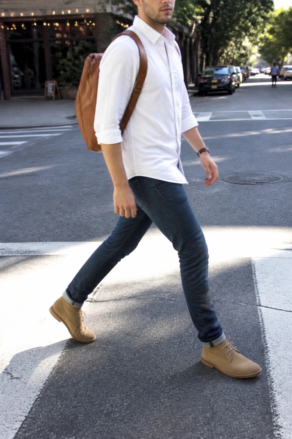 Man in white shirt and jeans with brown backpack crossing sunny city street in tan boots