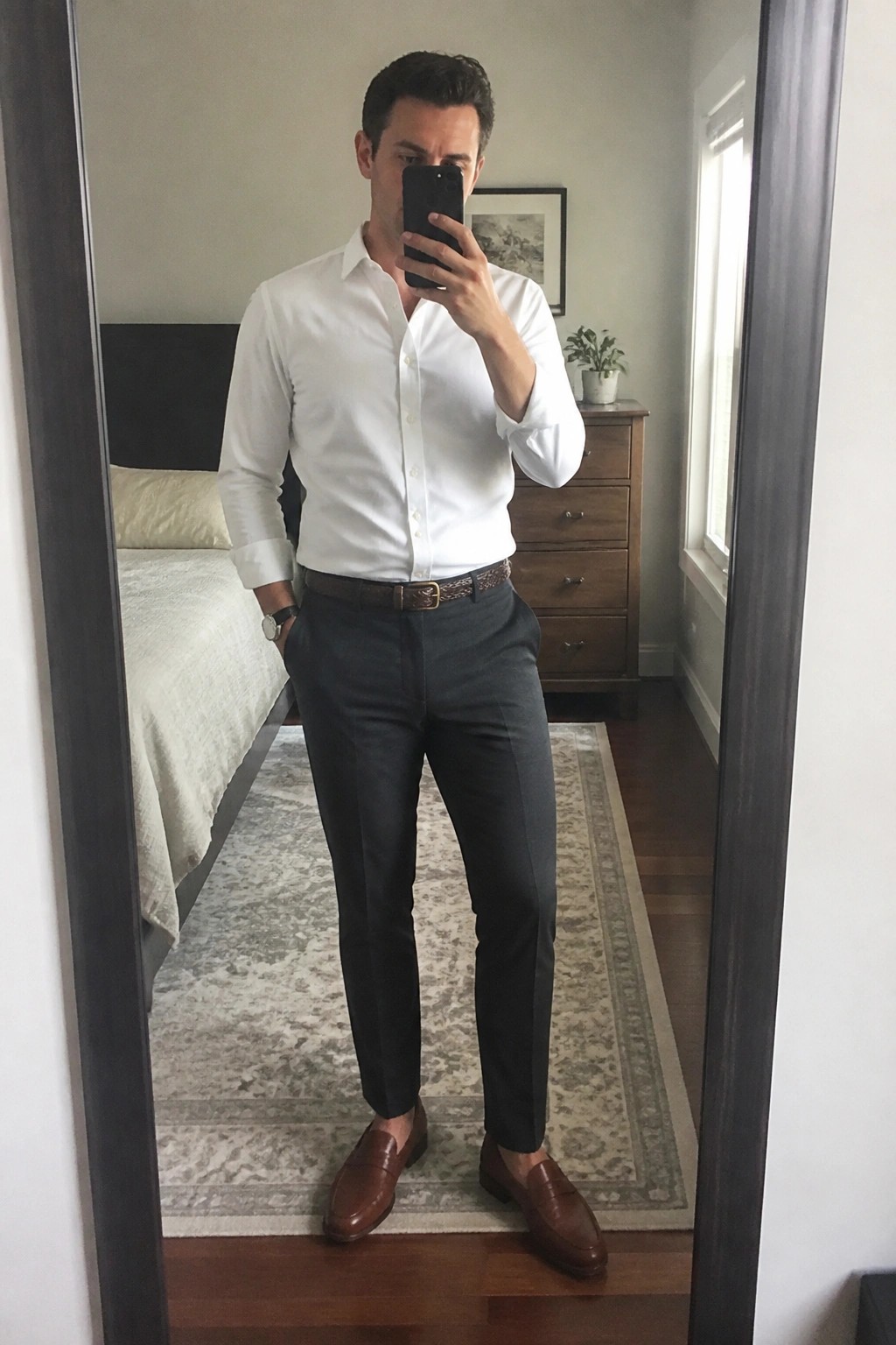 Man in white shirt and gray pants taking bedroom mirror selfie with phone.