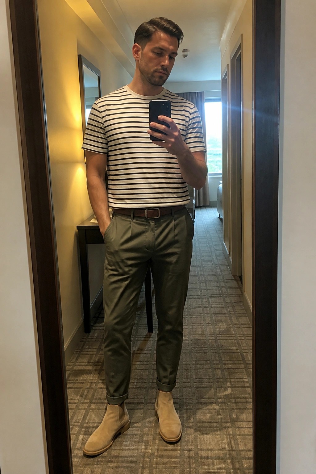 Man in striped shirt and olive pants taking mirror selfie in hallway