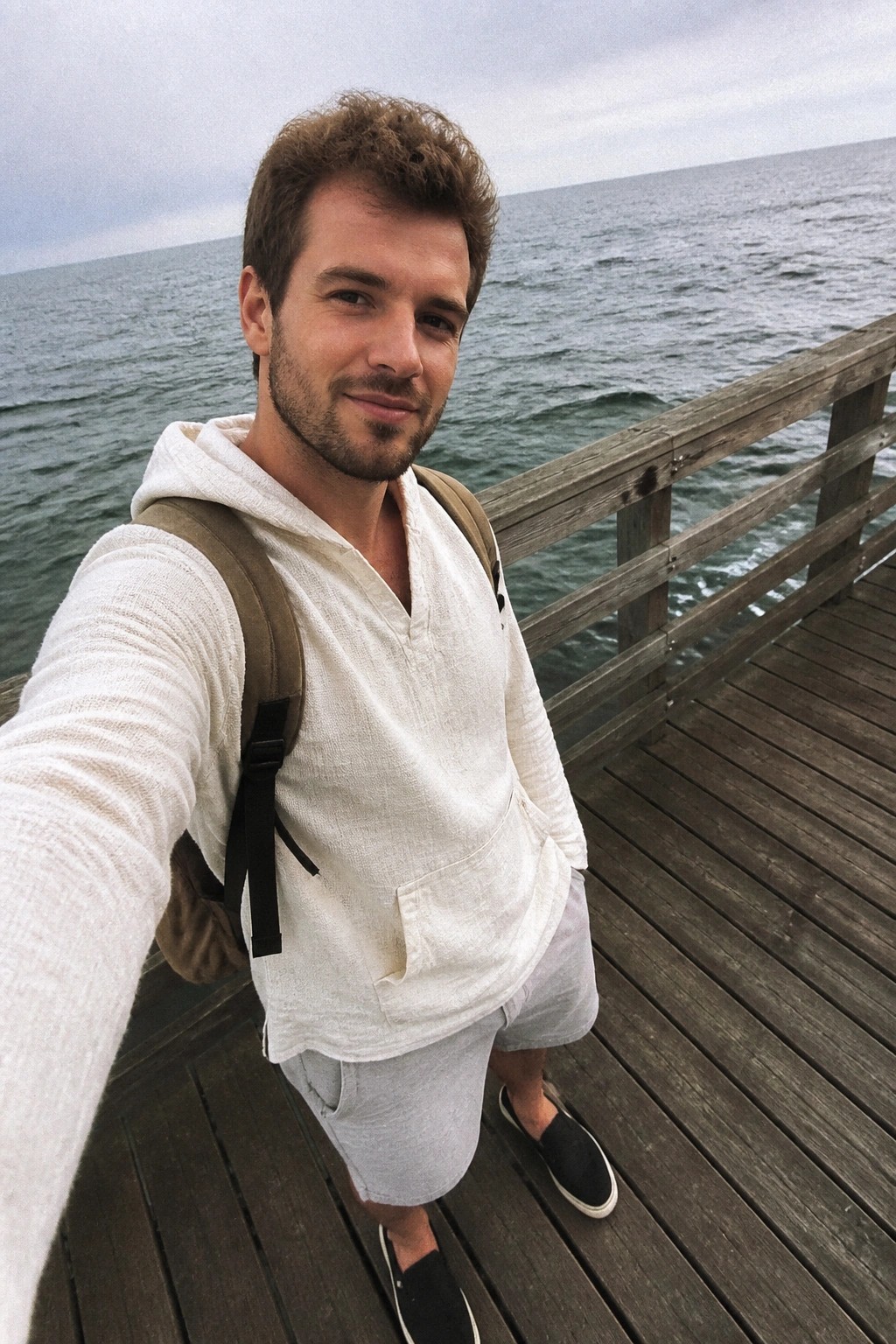 Man in white hooded shirt and gray shorts with backpack on ocean pier.