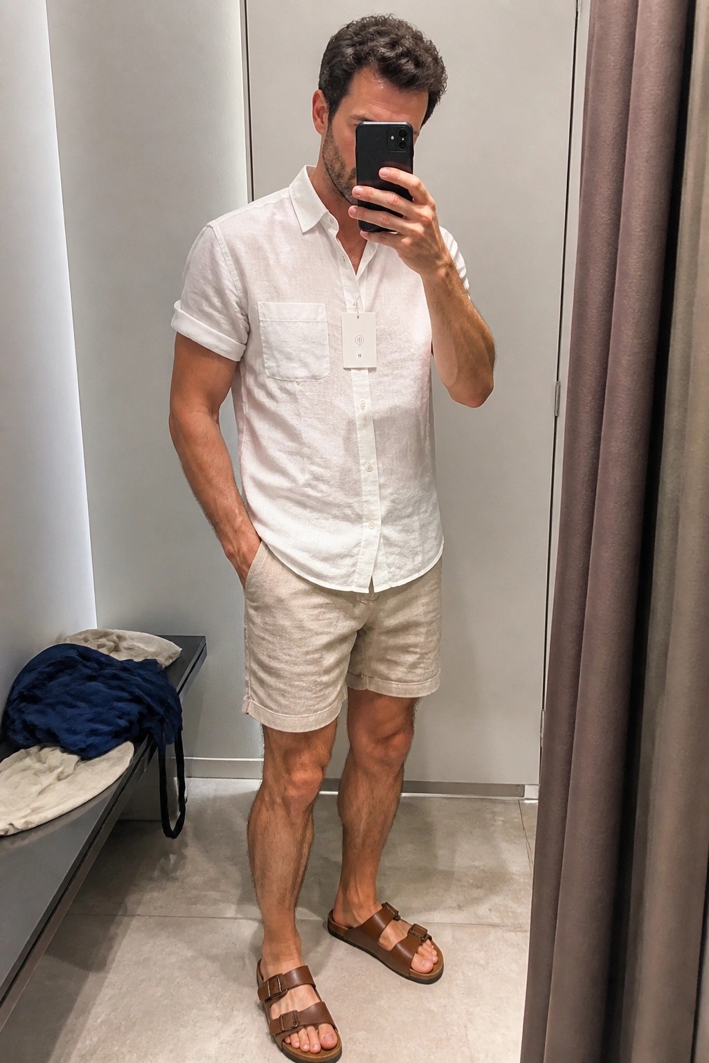 Man in white linen shirt and beige shorts taking mirror selfie in fitting room