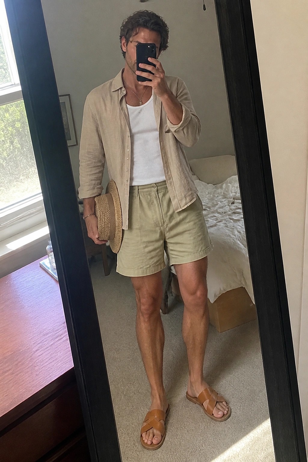 Man in beige linen shirt and green shorts taking mirror selfie with straw hat