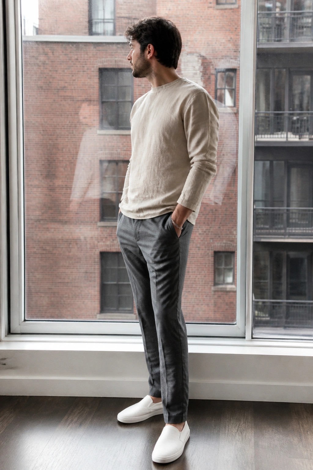 Man in beige long sleeve linen shirt tucked into gray slim pants with white slip on shoes and silver bracelet standing in front of large window.