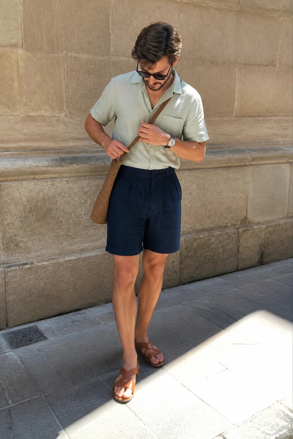 Man in light green shirt and navy shorts adjusting woven shoulder bag strap.