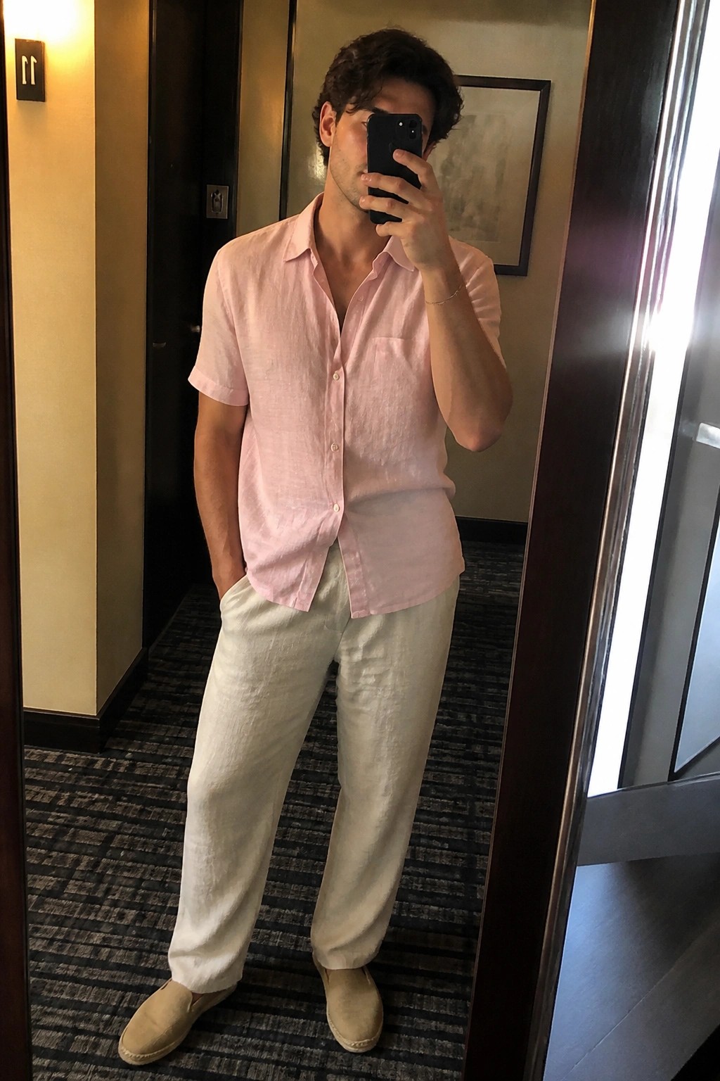 Man in pink linen shirt and white pants taking mirror selfie in hallway.